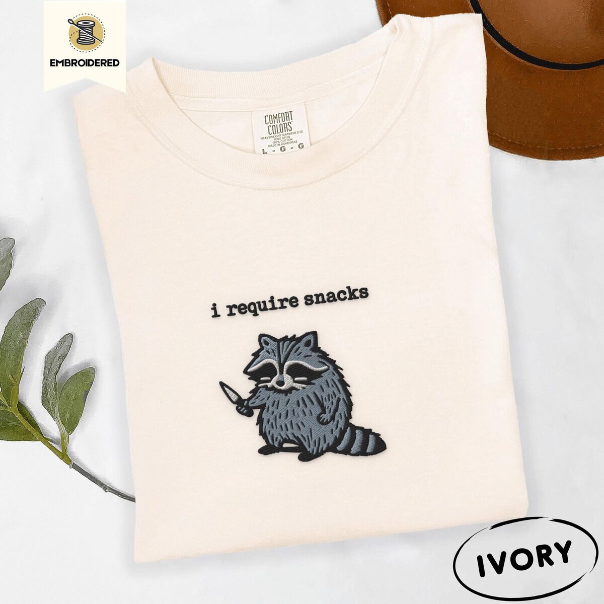 Raccoon Shirt – Ivory Ivory Comfort Colors T-shirt embroidered with a feral raccoon holding a little knife under the words i require snacks embroidered on the chest