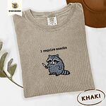 Khaki Comfort Colors T-shirt embroidered with a feral raccoon holding a little knife under the words i require snacks embroidered on the chest