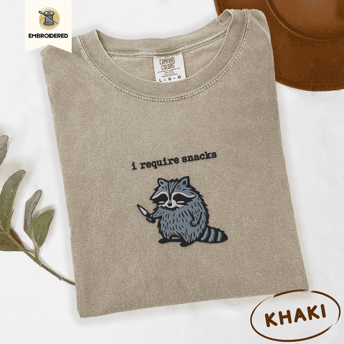 Raccoon Shirt – Khaki Khaki Comfort Colors T-shirt embroidered with a feral raccoon holding a little knife under the words i require snacks embroidered on the chest