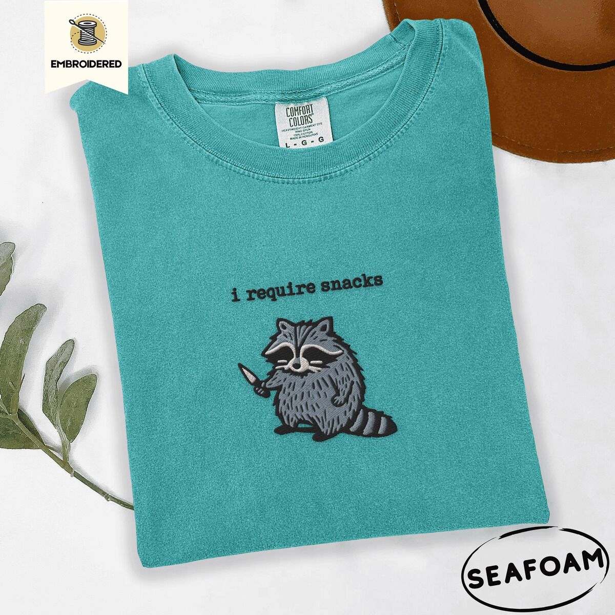 Raccoon Shirt – Seafoam Seafoam Comfort Colors T-shirt embroidered with a feral raccoon holding a little knife under the words i require snacks embroidered on the chest