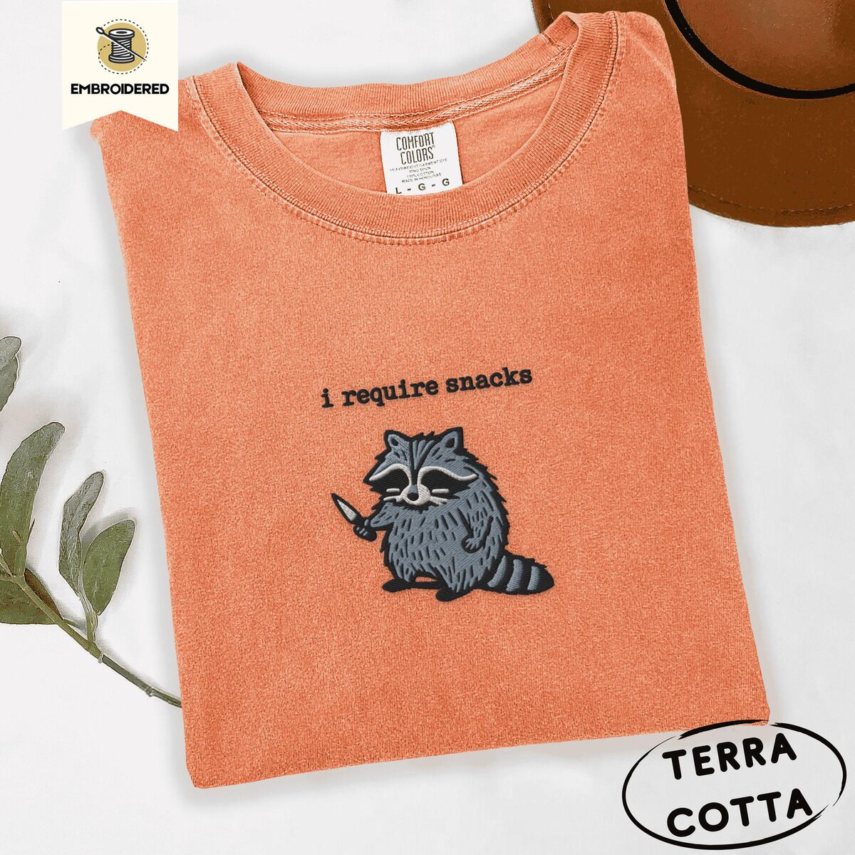 Raccoon Shirt – Terracotta Terracotta Comfort Colors T-shirt embroidered with a feral raccoon holding a little knife under the words i require snacks embroidered on the chest