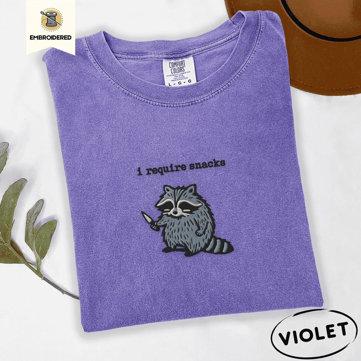 Raccoon Shirt – Violet Violet Comfort Colors T-shirt embroidered with a feral raccoon holding a little knife under the words i require snacks embroidered on the chest