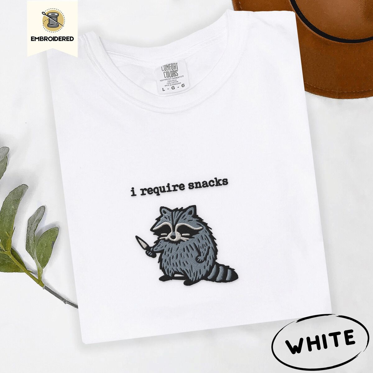 Raccoon Shirt – White White Comfort Colors T-shirt embroidered with a feral raccoon holding a little knife under the words i require snacks embroidered on the chest