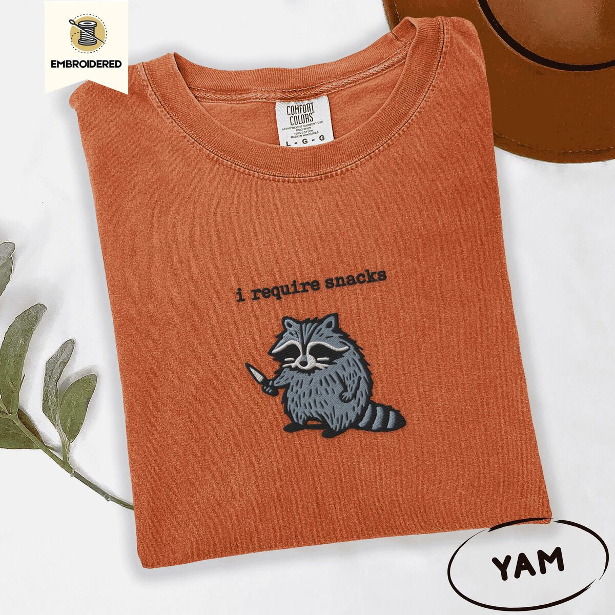 Raccoon Shirt – Yam Yam Comfort Colors T-shirt embroidered with a feral raccoon holding a little knife under the words i require snacks embroidered on the chest
