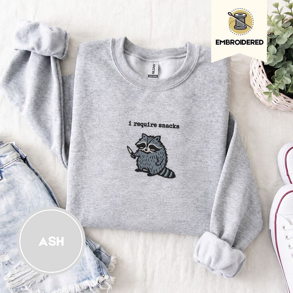 Ash crewneck sweatshirt folded with denim shorts, white sneakers and a potted plant, embroidered with a feral raccoon holding a little knife under the words i require snacks embroidered on the chest