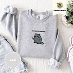 Ash crewneck sweatshirt folded with denim shorts, white sneakers and a potted plant, embroidered with a feral raccoon holding a little knife under the words i require snacks embroidered on the chest