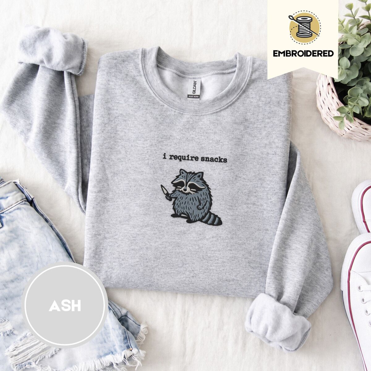 Raccoon Sweatshirt – Ash Ash crewneck sweatshirt folded with denim shorts, white sneakers and a potted plant, embroidered with a feral raccoon holding a little knife under the words i require snacks embroidered on the chest