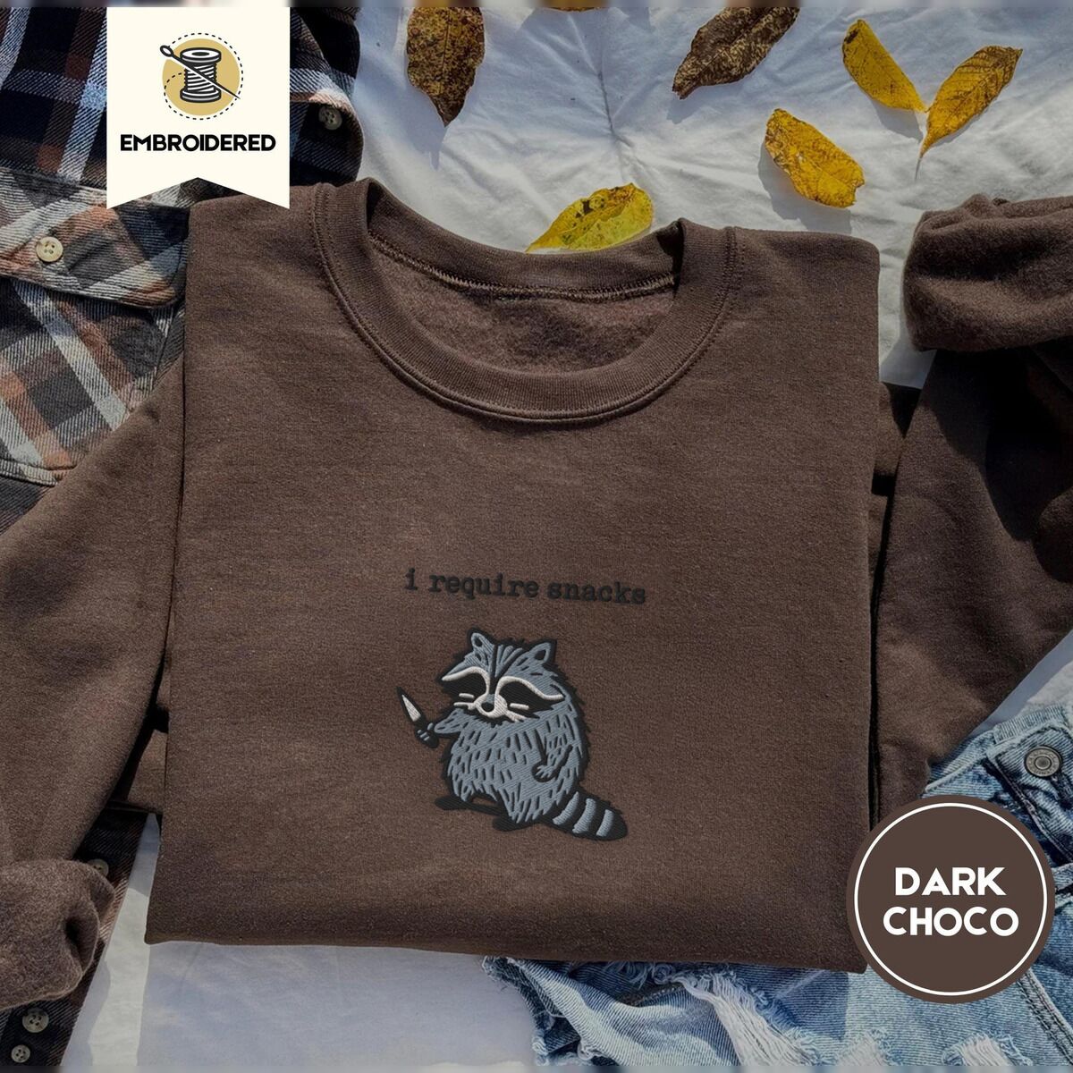Raccoon Sweatshirt – Dark Chocolate Dark Chocolate crewneck sweatshirt on a plaid flannel shirt with yellow autumn leaves and denim shorts, embroidered with a feral raccoon holding a little knife under the words i require snacks embroidered on the chest