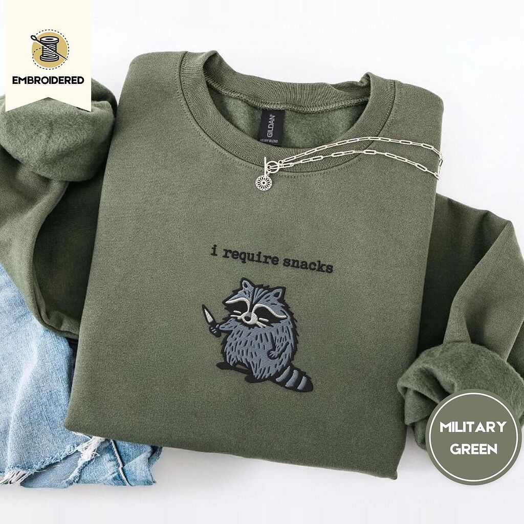 Military Green crewneck sweatshirt folded with light jeans and a silver chain necklace, embroidered with a feral raccoon holding a little knife under the words i require snacks embroidered on the chest