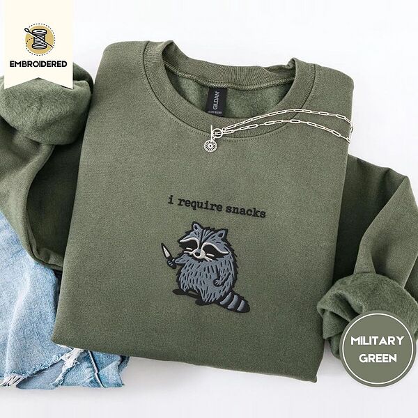 Military Green crewneck sweatshirt folded with light jeans and a silver chain necklace, embroidered with a feral raccoon holding a little knife under the words i require snacks embroidered on the chest