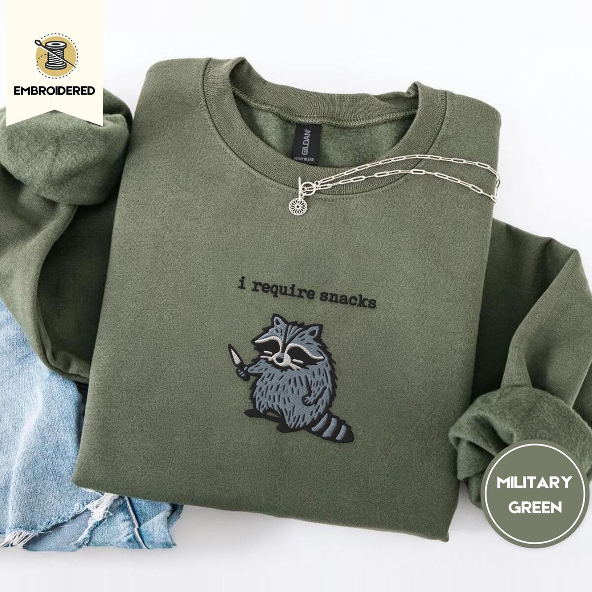 Raccoon Sweatshirt – Military Green Military Green crewneck sweatshirt folded with light jeans and a silver chain necklace, embroidered with a feral raccoon holding a little knife under the words i require snacks embroidered on the chest