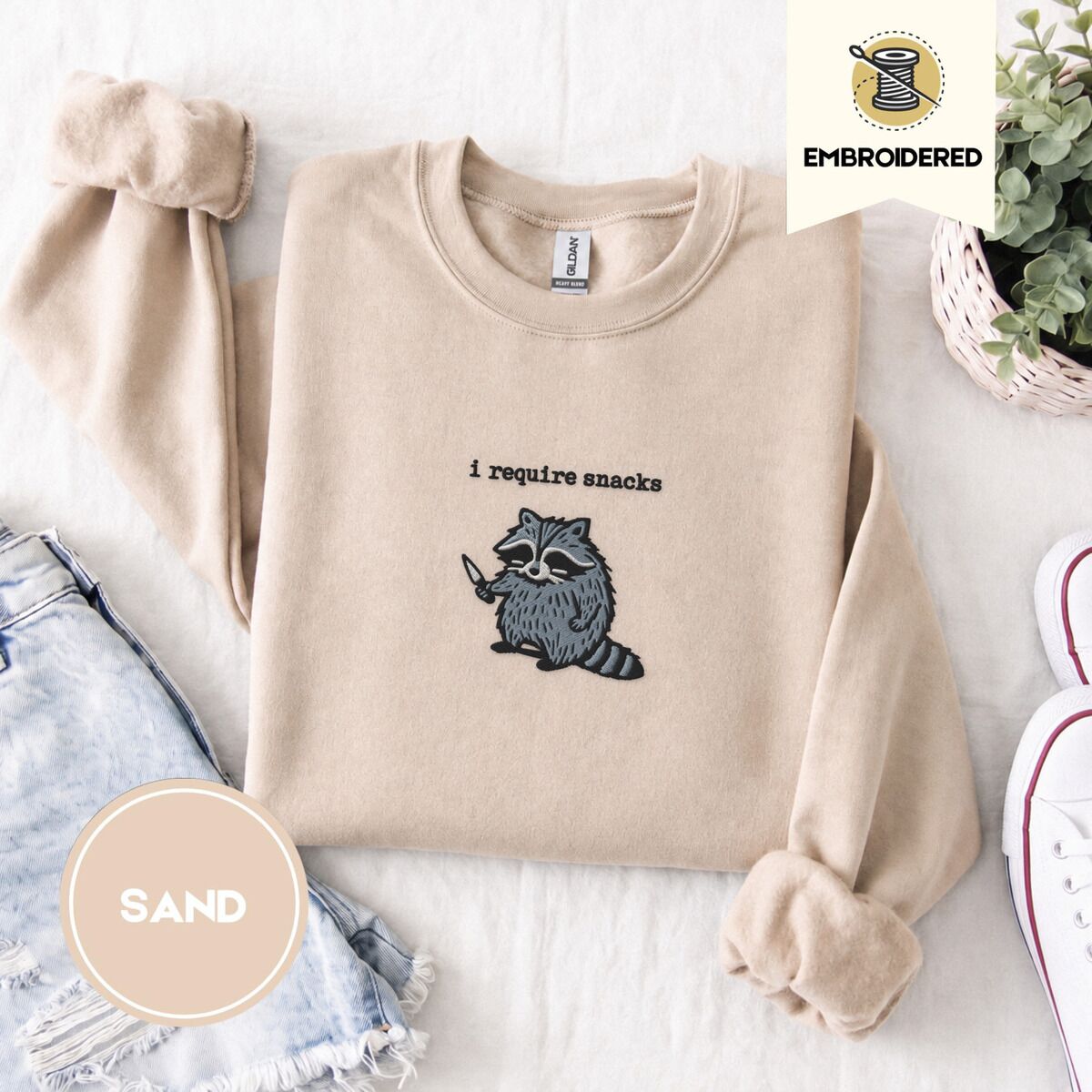 Raccoon Sweatshirt – Sand Sand crewneck sweatshirt folded with denim shorts, white sneakers and a potted plant, embroidered with a feral raccoon holding a little knife under the words i require snacks embroidered on the chest