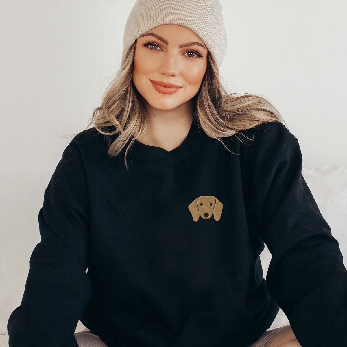Red Dachshund Sweatshirt – Black Woman in a beanie wearing a Black crewneck sweatshirt embroidered with a red dachshund face with long floppy ears, round black eyes and a little smile on the left chest