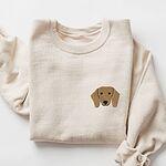 Sand crewneck sweatshirt laid flat on white, embroidered with a red dachshund face with long floppy ears, round black eyes and a little smile on the left chest