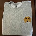 Sport Grey crewneck sweatshirt folded on a dark wood table, embroidered with a red dachshund face with long floppy ears, round black eyes and a little smile on the left chest