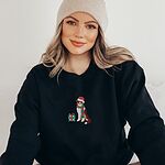 Woman in a beanie sitting in a Black crewneck sweatshirt embroidered with a red tri Australian shepherd in a Santa hat and green and red striped scarf beside a green polka dot gift box