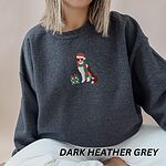 Woman sitting on a bed in a Dark Heather crewneck sweatshirt embroidered with a red tri Australian shepherd in a Santa hat and green and red striped scarf beside a green polka dot gift box