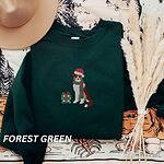 Forest Green crewneck sweatshirt laid flat with a tan hat and dried pampas grass, embroidered with a red tri Australian shepherd in a Santa hat and green and red striped scarf beside a green polka dot gift box