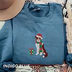 Indigo Blue crewneck sweatshirt laid flat with a tan hat, embroidered with a red tri Australian shepherd in a Santa hat and green and red striped scarf beside a green polka dot gift box