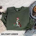 Military Green crewneck sweatshirt laid flat on a white fur rug with pine cones and a knit blanket, embroidered with a red tri Australian shepherd in a Santa hat and green and red striped scarf beside a green polka dot gift box