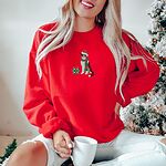Woman with a mug by a Christmas tree in a Red crewneck sweatshirt embroidered with a red tri Australian shepherd in a Santa hat and green and red striped scarf beside a green polka dot gift box