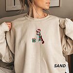 Woman pulling at the collar of a sand crewneck sweatshirt embroidered with a red tri Australian shepherd in a Santa hat and green and red striped scarf beside a green polka dot gift box