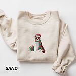 Sand crewneck sweatshirt laid flat on a white background, embroidered with a red tri Australian shepherd in a Santa hat and green and red striped scarf beside a green polka dot gift box