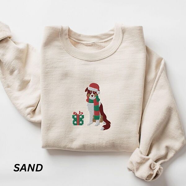 Sand crewneck sweatshirt laid flat on a white background, embroidered with a red tri Australian shepherd in a Santa hat and green and red striped scarf beside a green polka dot gift box