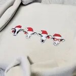 Close-up of four embroidered Santa cows on a Sand crewneck, a Holstein, two cows with red bows and one in red sunglasses