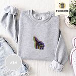 Ash crewneck sweatshirt folded with denim shorts, white sneakers and a potted plant, embroidered with a startled black cat with an arched back, a puffed tail and yellow eyes, outlined in layered rainbow neon stitches