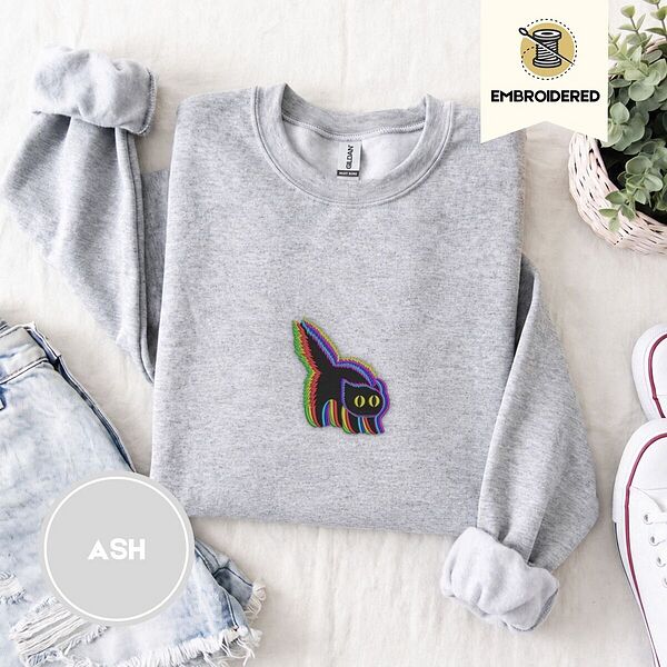 Ash crewneck sweatshirt folded with denim shorts, white sneakers and a potted plant, embroidered with a startled black cat with an arched back, a puffed tail and yellow eyes, outlined in layered rainbow neon stitches