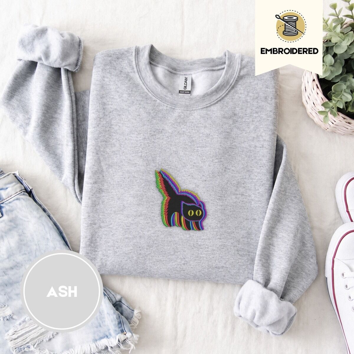 Scared Black Cat Sweatshirt – Ash Ash crewneck sweatshirt folded with denim shorts, white sneakers and a potted plant, embroidered with a startled black cat with an arched back, a puffed tail and yellow eyes, outlined in layered rainbow neon stitches