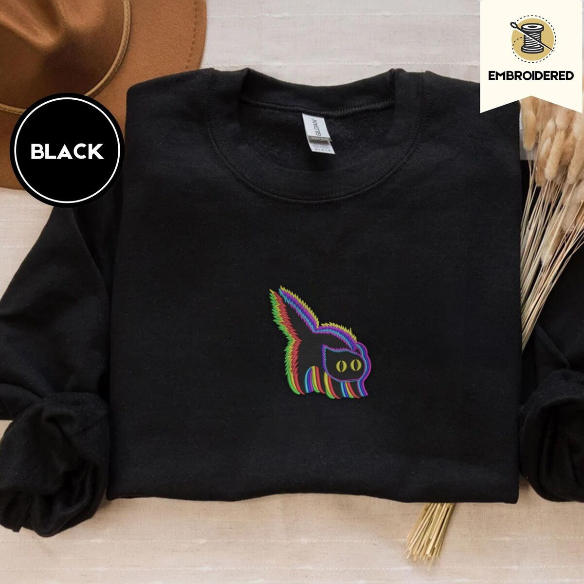 Scared Black Cat Sweatshirt – Black Black crewneck sweatshirt with a brown felt hat and dried bunny tail grass, embroidered with a startled black cat with an arched back, a puffed tail and yellow eyes, outlined in layered rainbow neon stitches