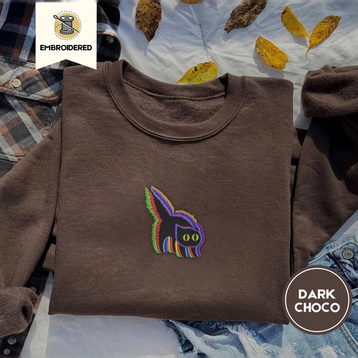 Scared Black Cat Sweatshirt – Dark Chocolate Dark Chocolate crewneck sweatshirt on a plaid flannel shirt with yellow autumn leaves and denim shorts, embroidered with a startled black cat with an arched back, a puffed tail and yellow eyes, outlined in layered rainbow neon stitches