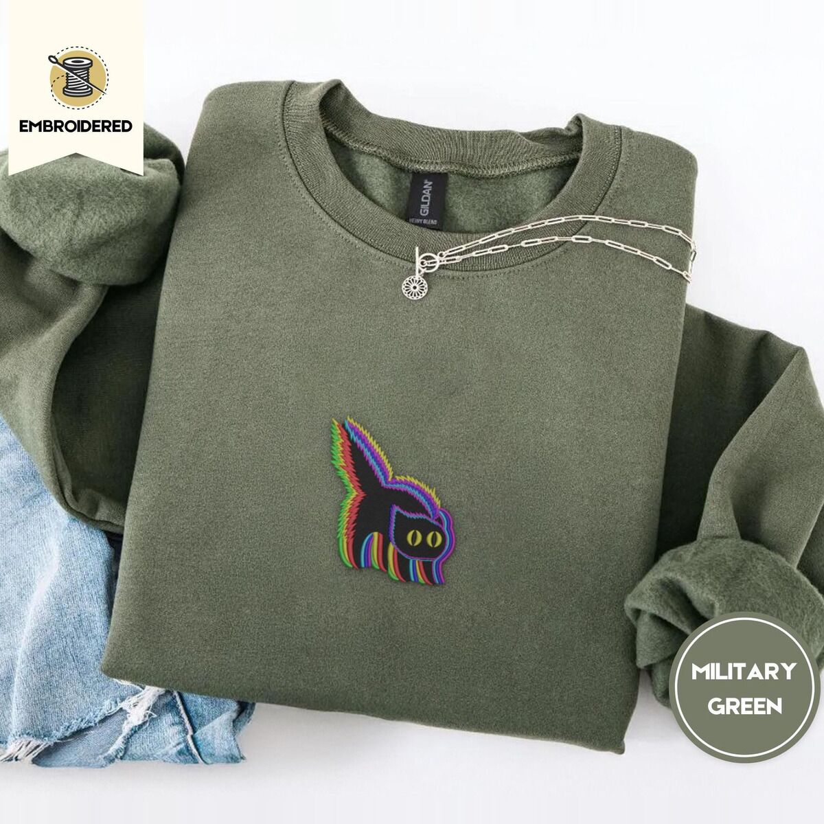 Scared Black Cat Sweatshirt – Military Green Military Green crewneck sweatshirt folded with light jeans and a silver chain necklace, embroidered with a startled black cat with an arched back, a puffed tail and yellow eyes, outlined in layered rainbow neon stitches