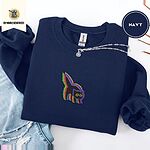 Navy crewneck sweatshirt folded with ripped jeans and a silver chain necklace, embroidered with a startled black cat with an arched back, a puffed tail and yellow eyes, outlined in layered rainbow neon stitches
