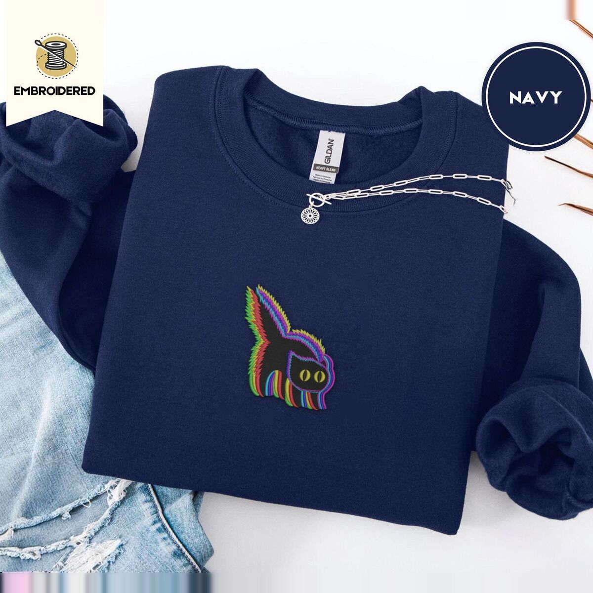Scared Black Cat Sweatshirt – Navy Navy crewneck sweatshirt folded with ripped jeans and a silver chain necklace, embroidered with a startled black cat with an arched back, a puffed tail and yellow eyes, outlined in layered rainbow neon stitches
