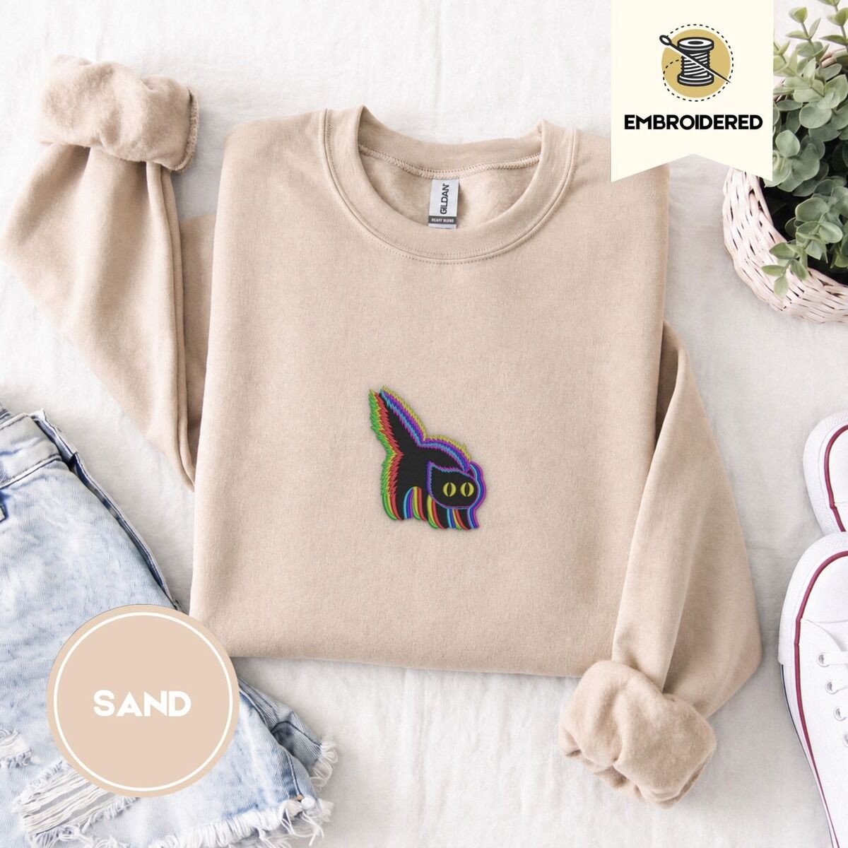 Scared Black Cat Sweatshirt – Sand Sand crewneck sweatshirt folded with denim shorts, white sneakers and a potted plant, embroidered with a startled black cat with an arched back, a puffed tail and yellow eyes, outlined in layered rainbow neon stitches