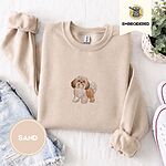 Sand crewneck sweatshirt folded with denim shorts, white sneakers and a potted plant, embroidered with a gold and white Shih Tzu puppy with a long silky coat and a plumed tail