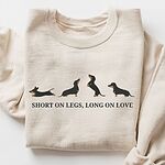 Sand crewneck sweatshirt laid flat on white, embroidered with four black and tan dachshunds in a row above the words SHORT ON LEGS, LONG ON LOVE across the chest