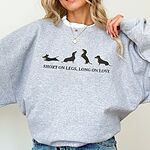 Woman in an oversized grey crewneck sweatshirt and jeans, embroidered with four black and tan dachshunds in a row above the words SHORT ON LEGS, LONG ON LOVE across the chest