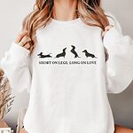 Woman in a White crewneck sweatshirt embroidered with four black and tan dachshunds in a row above the words SHORT ON LEGS, LONG ON LOVE across the chest
