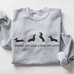 Sport Grey crewneck sweatshirt laid flat on white, embroidered with four black and tan dachshunds in a row above the words SHORT ON LEGS, LONG ON LOVE across the chest