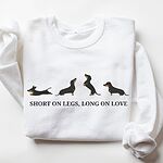 White crewneck sweatshirt laid flat on white, embroidered with four black and tan dachshunds in a row above the words SHORT ON LEGS, LONG ON LOVE across the chest