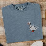 Ice Blue T-shirt laid flat on wood beside a white sneaker, embroidered with a white goose and the words silly dada in white thread