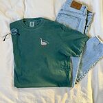 Green T-shirt laid flat on a bed with jeans and glasses, embroidered with a small white goose and the words silly dada