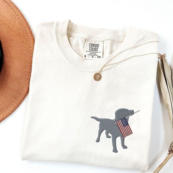 Ivory T-shirt folded beside a hat with a silver lab silhouette carrying an American flag on the left chest