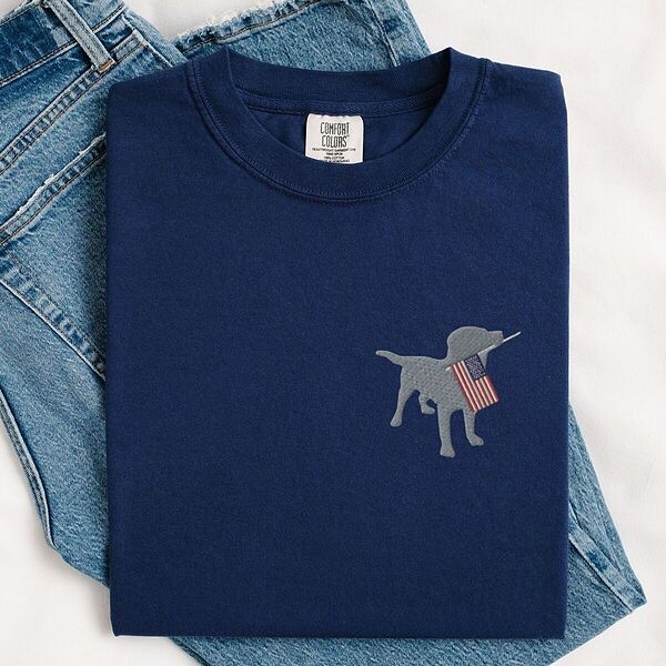 True Navy T-shirt folded on blue jeans, a gray Labrador holding a stars-and-stripes flag stitched on the left chest