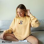 Smiling woman on a sofa in a Butter crewneck sweatshirt embroidered with a silver lab in a white ghost sheet holding a jack-o'-lantern bucket