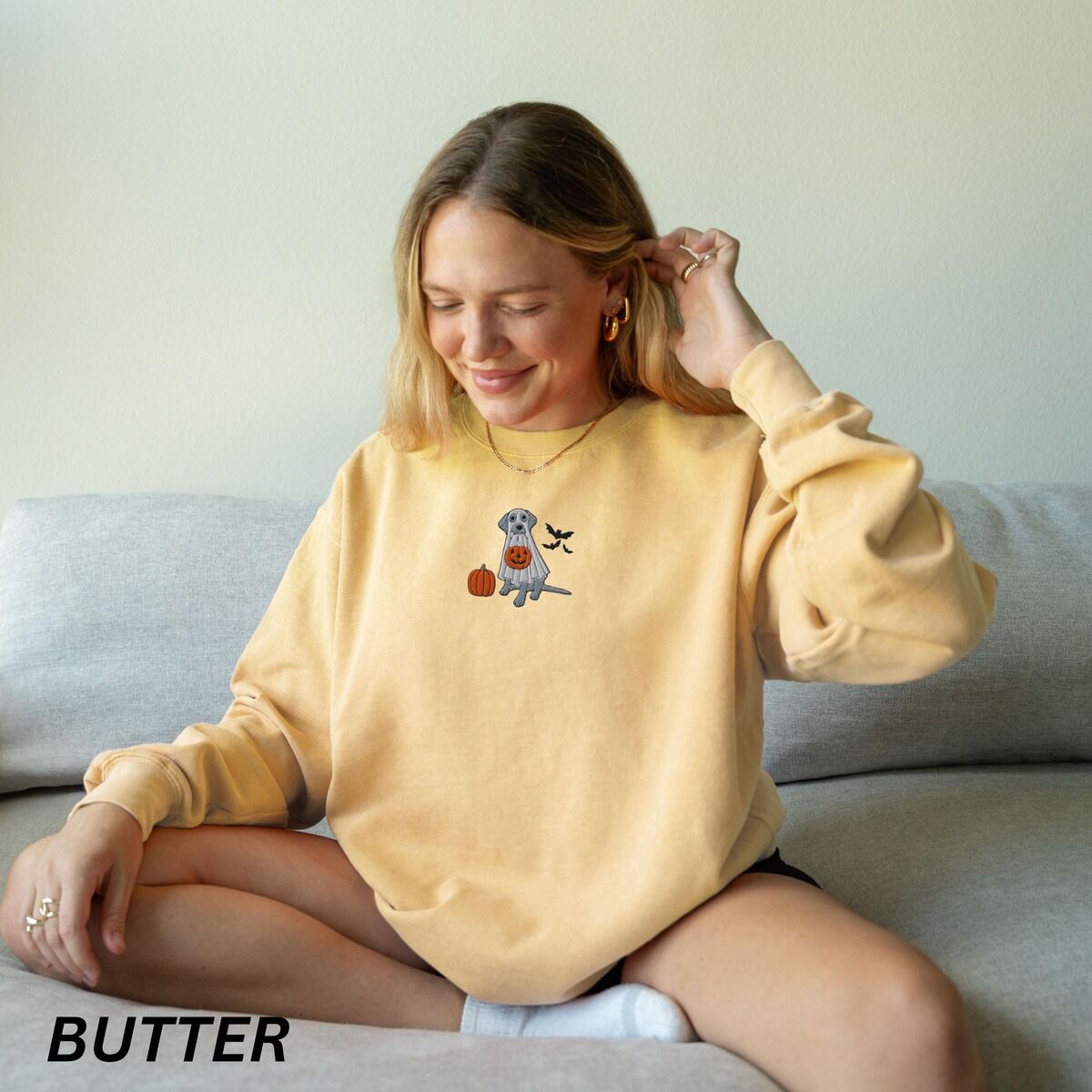 Silver Lab Ghost Sweatshirt – Butter Smiling woman on a sofa in a Butter crewneck sweatshirt embroidered with a silver lab in a white ghost sheet holding a jack-o'-lantern bucket